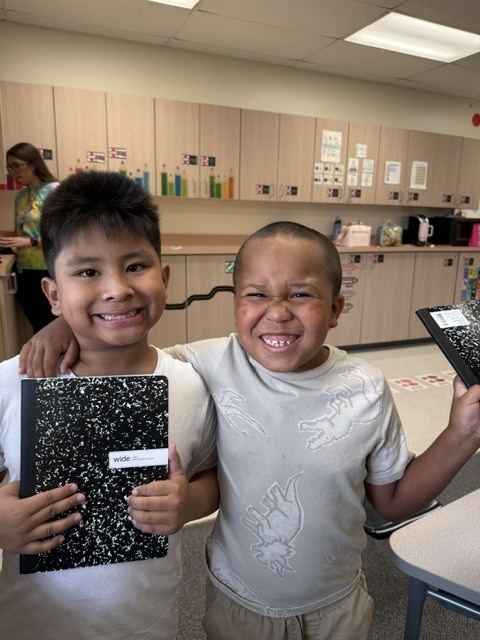 Two young boys in a classroom hold up books, smiling. A wooden counter and cabinets are behind them.