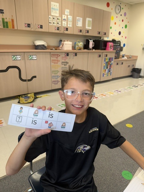 A child in a classroom holds a card with letters and numbers. They wear glasses. The room has cabinets and colorful decorations.