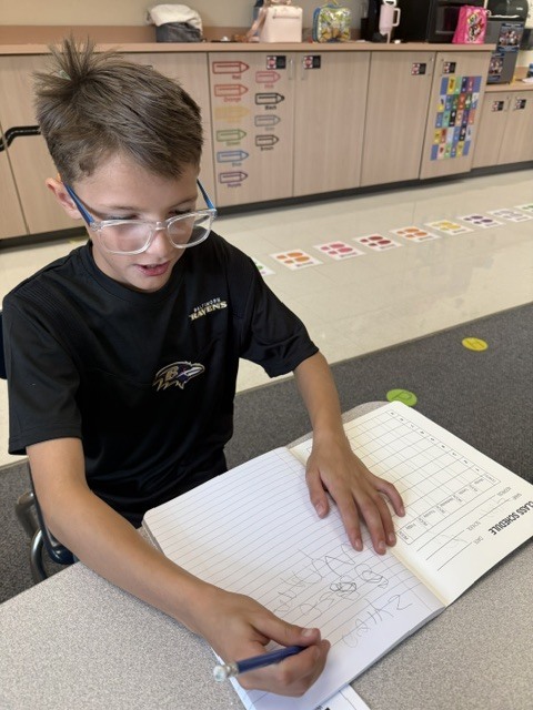 A student with glasses sits at a table, working on a notebook with a pen.