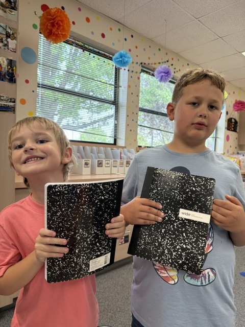 Two children in a room with books. One holds a black notebook and the other holds a notebook.