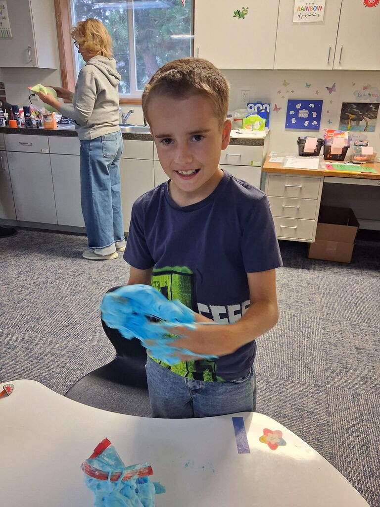 A young boy holding blue slime while smiling, with a woman behind him cleaning a countertop.