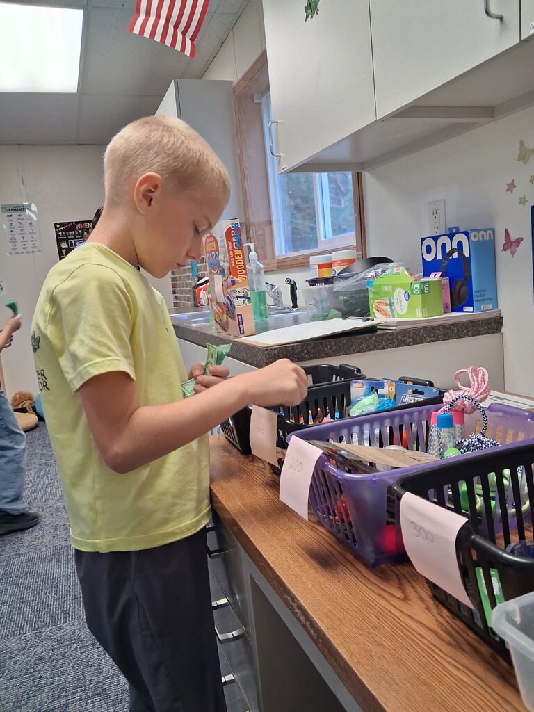 A boy in a yellow shirt arranges items in baskets. The room has white walls, a flag, and storage cabinets.