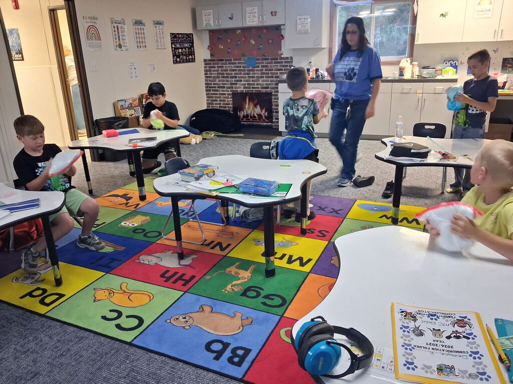 A classroom with children seated at tables. A teacher stands near a fireplace in the background. The floor has a colorful rug with alphabet letters.