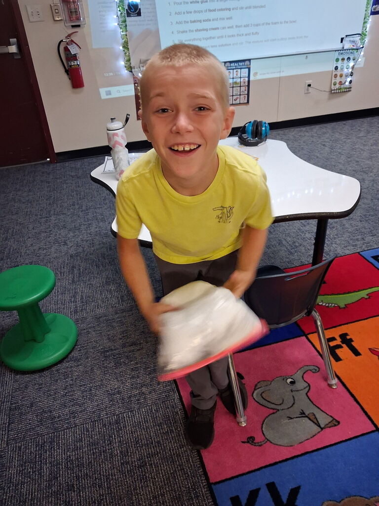 A child in a yellow t-shirt holds a rolled object while standing on a multicolored mat in a classroom.