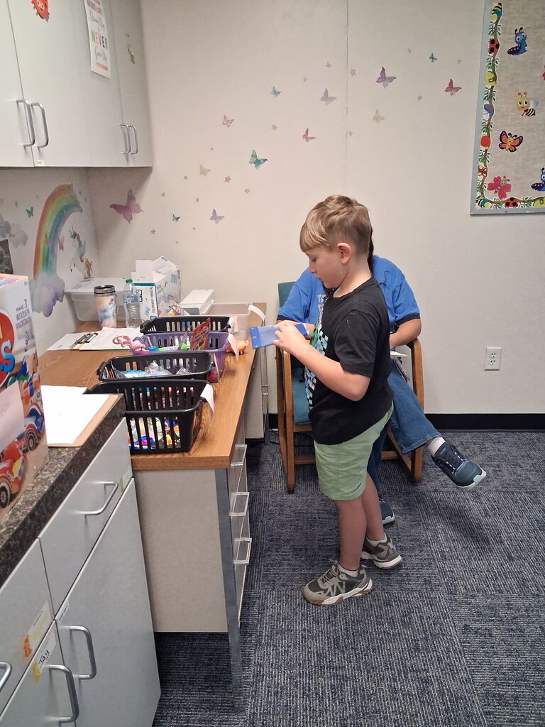 A young boy stands at a counter, holding a ribbon, next to an adult seated on a chair.