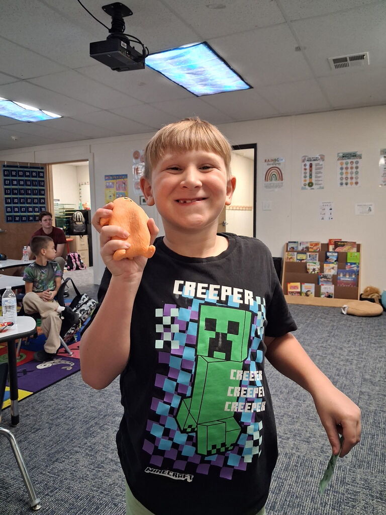 A boy in a black shirt holds a pink object, smiling at the camera. A projector and white ceiling lights are above.