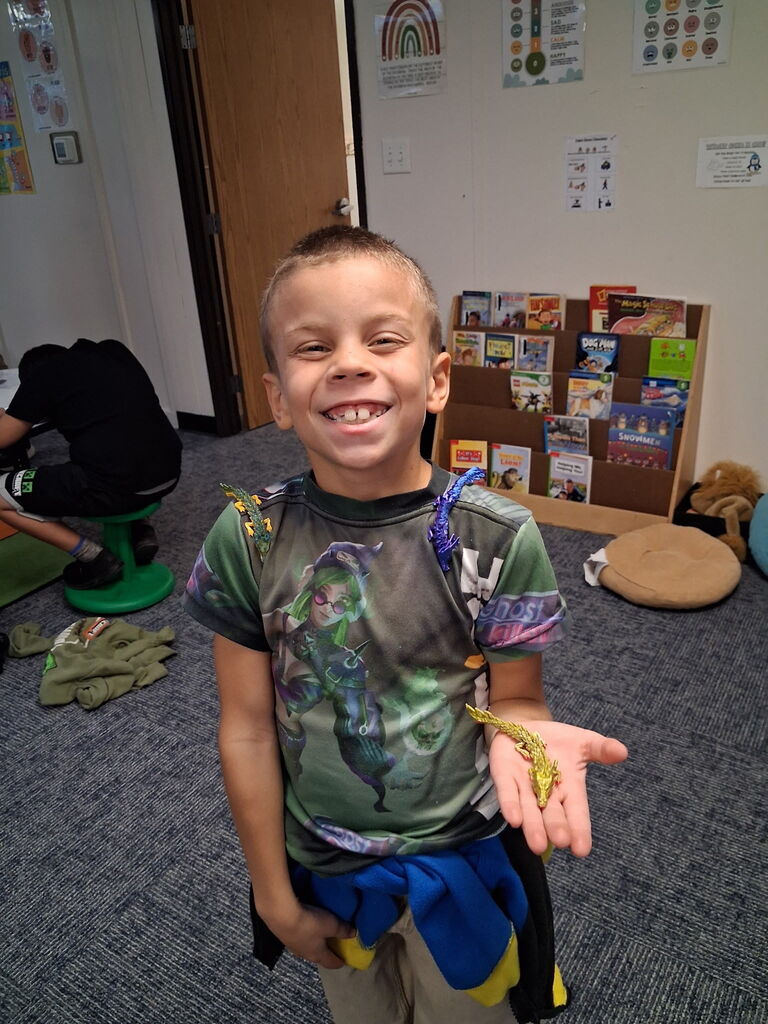 Child in classroom, smiling widely, wearing a green t-shirt with a colorful design. Bookshelves with books in background.