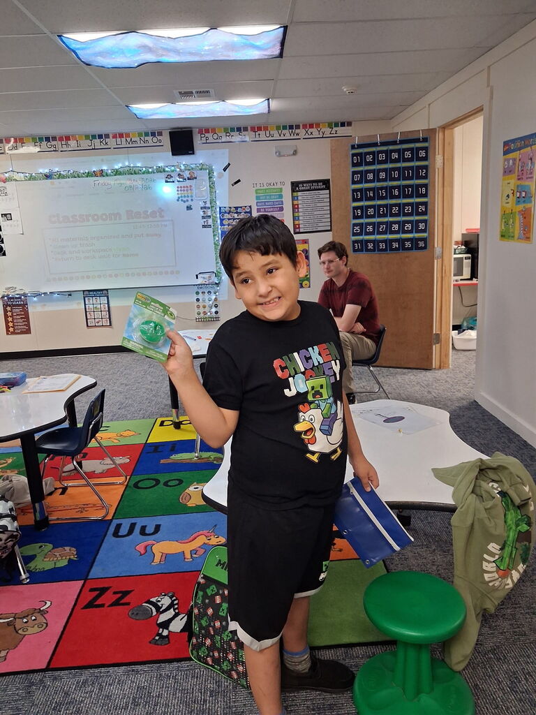 A boy holds a book, smiling, in a classroom. A man sits in the background. A colorful rug and desks are present.