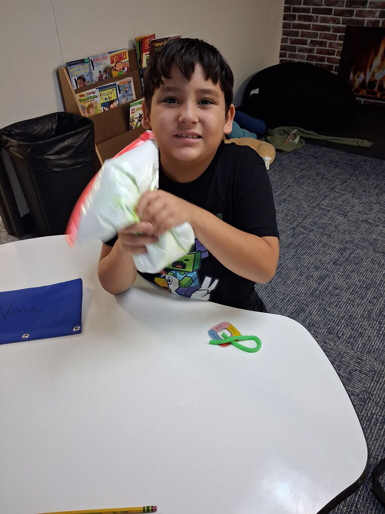 A boy seated at a table holds a folded towel, smiling. Items on the table include a blue folder and a green rubber band.
