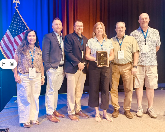 Six people stand on a stage, smiling, holding a plaque, with an American flag on the left.