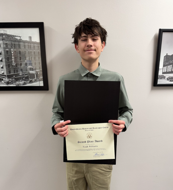 A young man in a green shirt holding a certificate in a black frame. Behind him, two framed black-and-white photographs of buildings.