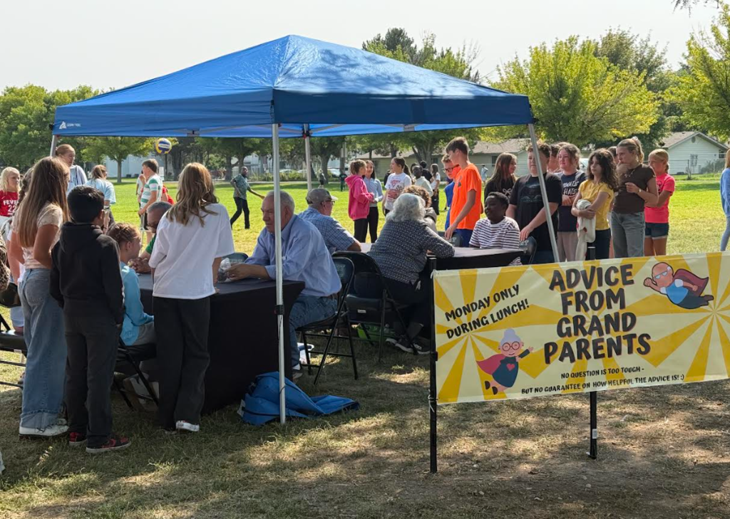 picture shows a blue canopy with students surrounding the tables. There is a yellow sign in front of the canopy.