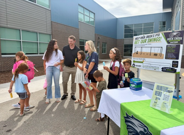 Adults and children stand in a group near a school building. They stand around a table with a poster.