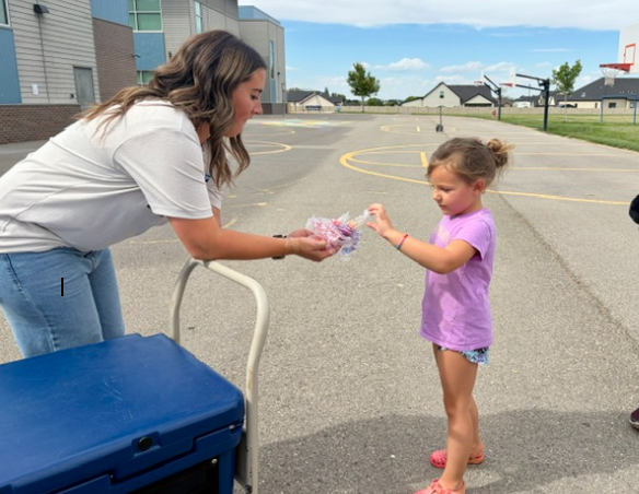 A woman hands a wrapped item to a girl, outside near a basketball court.
