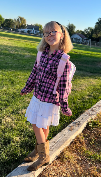 A young girl wearing glasses, a pink plaid long-sleeve shirt, a white skirt, and brown boots stands on a railroad tie in a grassy field.