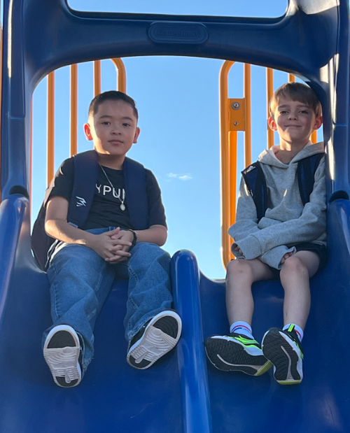 Two boys sit on a blue slide in an outdoor playground.