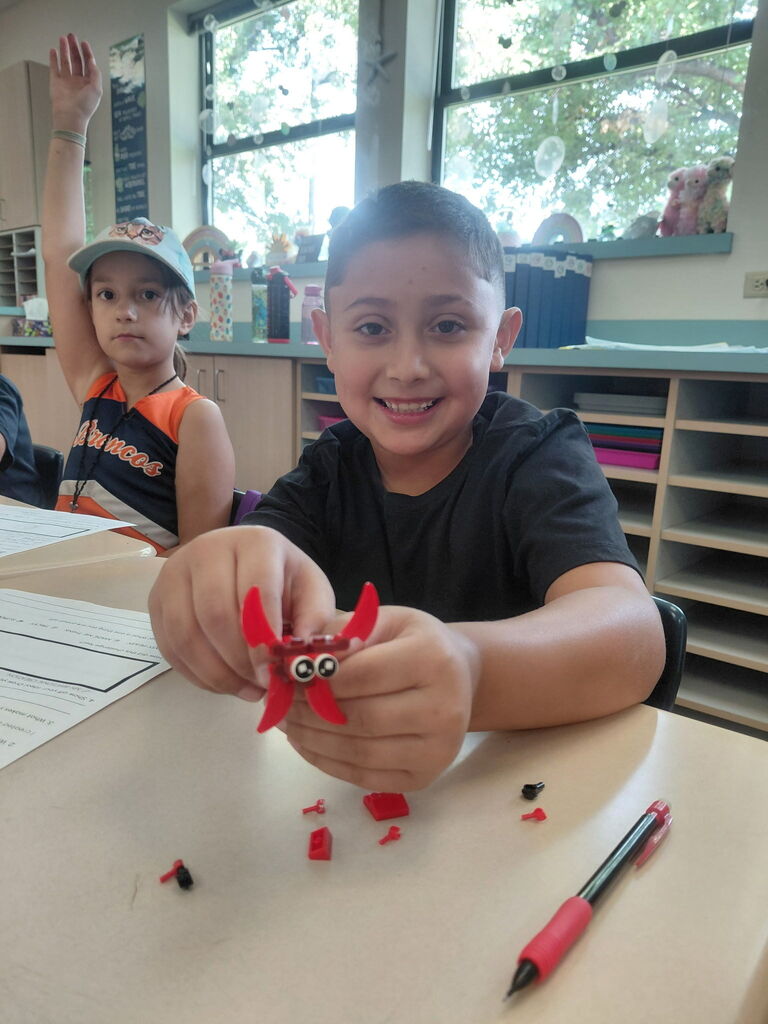 A child smiles, holding a red paper creation with googly eyes. Behind, another child raises a hand. A table, shelves, and windows are visible.