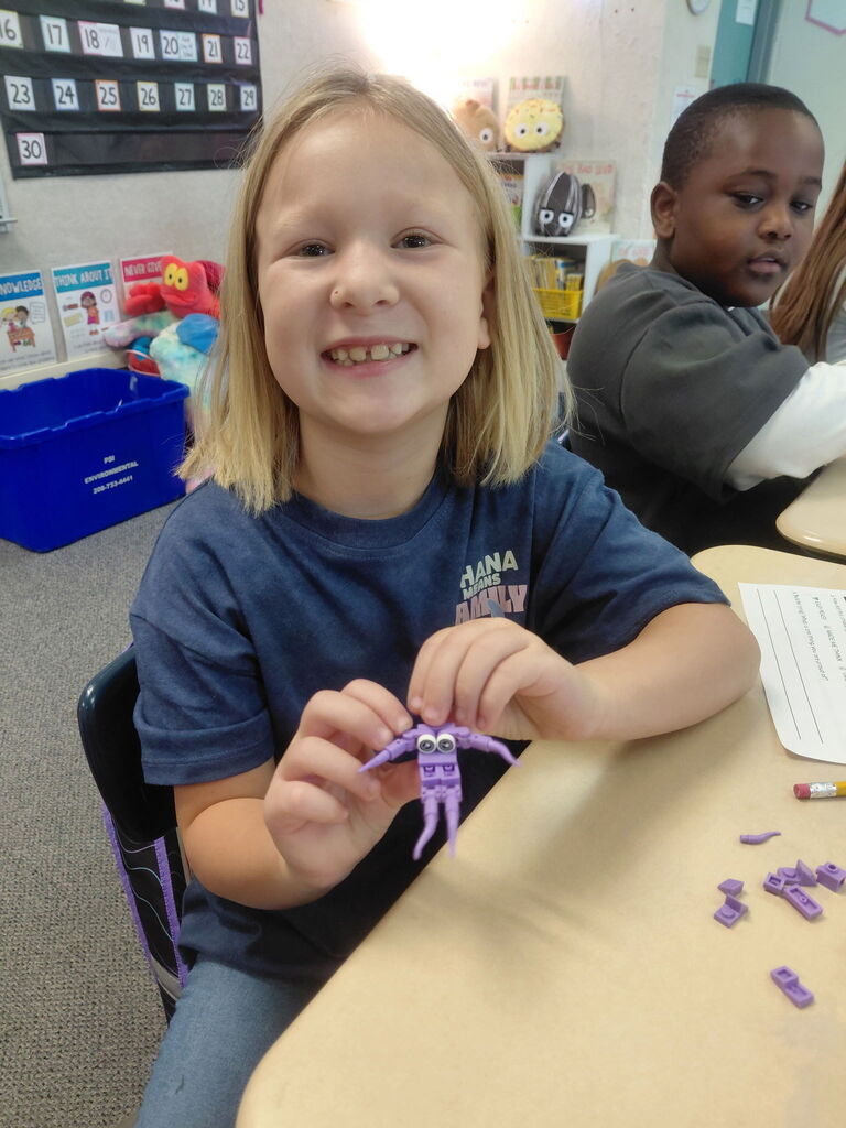 A child in a blue shirt smiles while holding a purple craft. Another child sits behind.