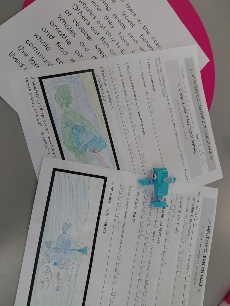 A desk displays a worksheet with blue drawings and text. A blue toy is placed between the papers.