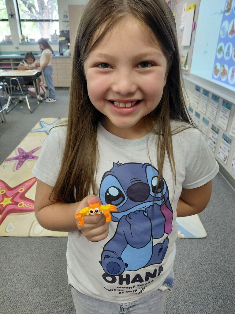 A young girl in a classroom holds a yellow toy. She wears a white shirt with a blue cartoon character.