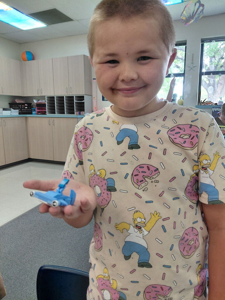 Child in a donut-themed shirt holding a blue toy airplane. Background shows a classroom with cabinets and windows.