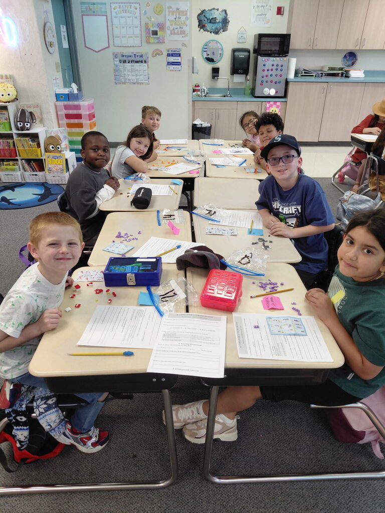 A classroom with children seated at desks. Papers, pens, and other school supplies are on the desks.