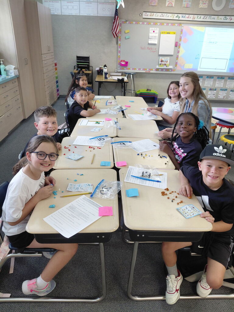 Children sit at desks, working on activities, with a teacher in the background, in a classroom.