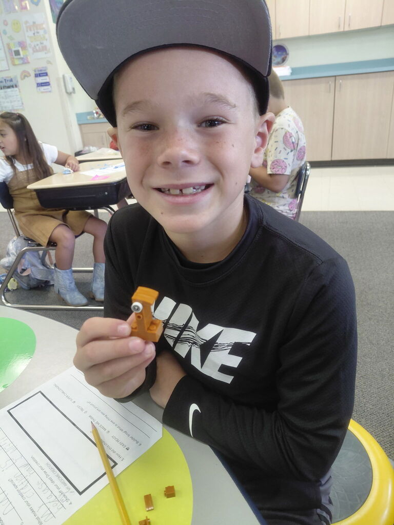 A boy in a black hat and shirt holds a small orange object in a classroom setting.
