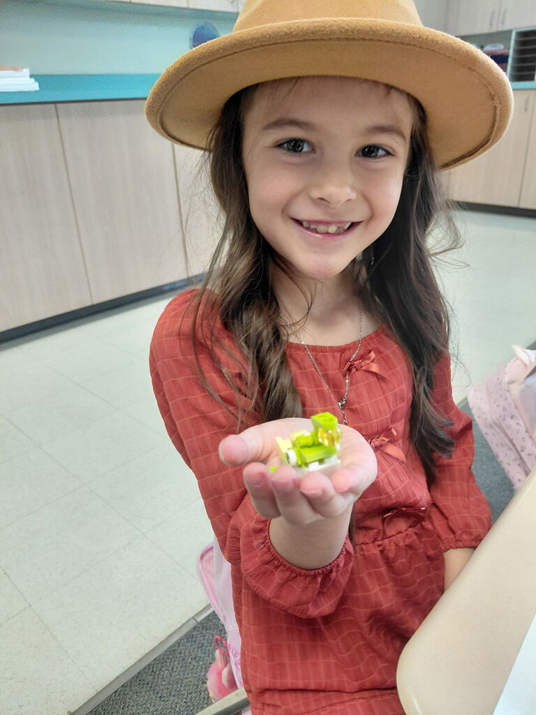 A girl in a red dress and hat holds a green vegetable in her hand, smiling.