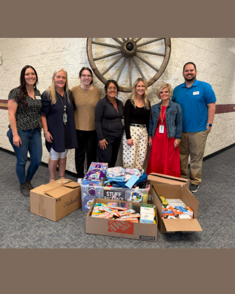 Six adults stand around three boxes of toys and two clear plastic bins filled with items. A large wooden wagon wheel is on the wall behind them.