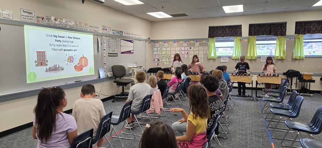 Inside a classroom, students sit on chairs facing a presentation on a projector screen. Some students wear sneakers.