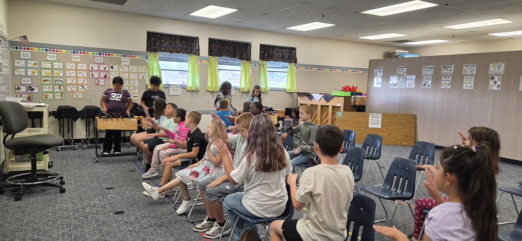 Group of students seated in chairs facing a teacher in a classroom with bulletin boards and chairs.