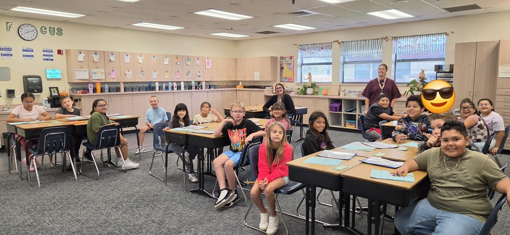 A classroom with students sitting at desks, a teacher, and a yellow emoji on a whiteboard.