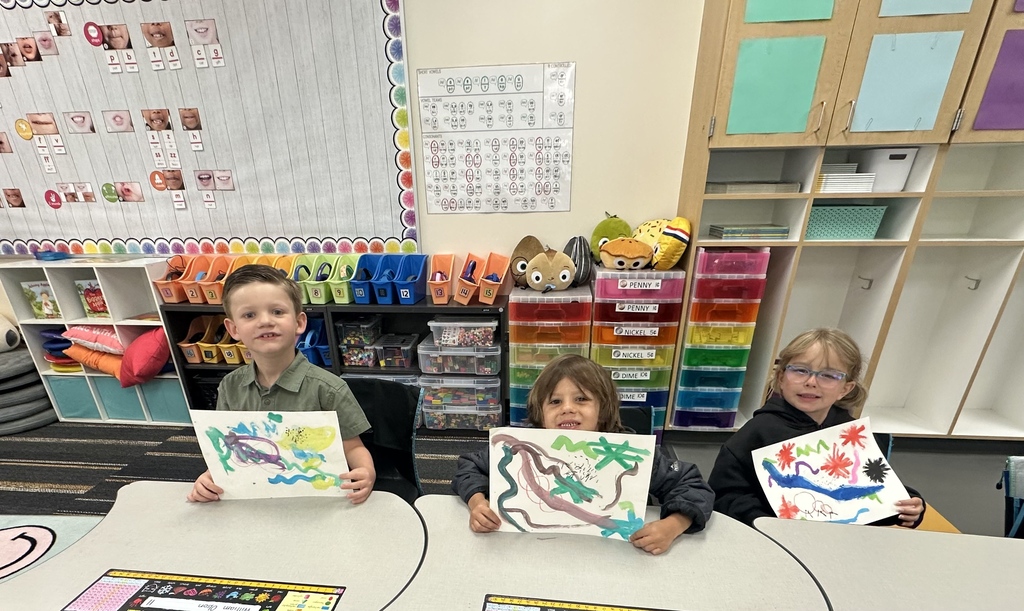 Three children hold up drawings of jungles and dinosaurs in a classroom with colorful shelves.