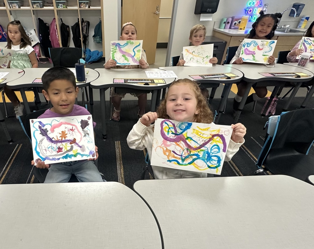 Children at desks in a classroom, holding colorful artwork. A boy and girl in front are smiling.
