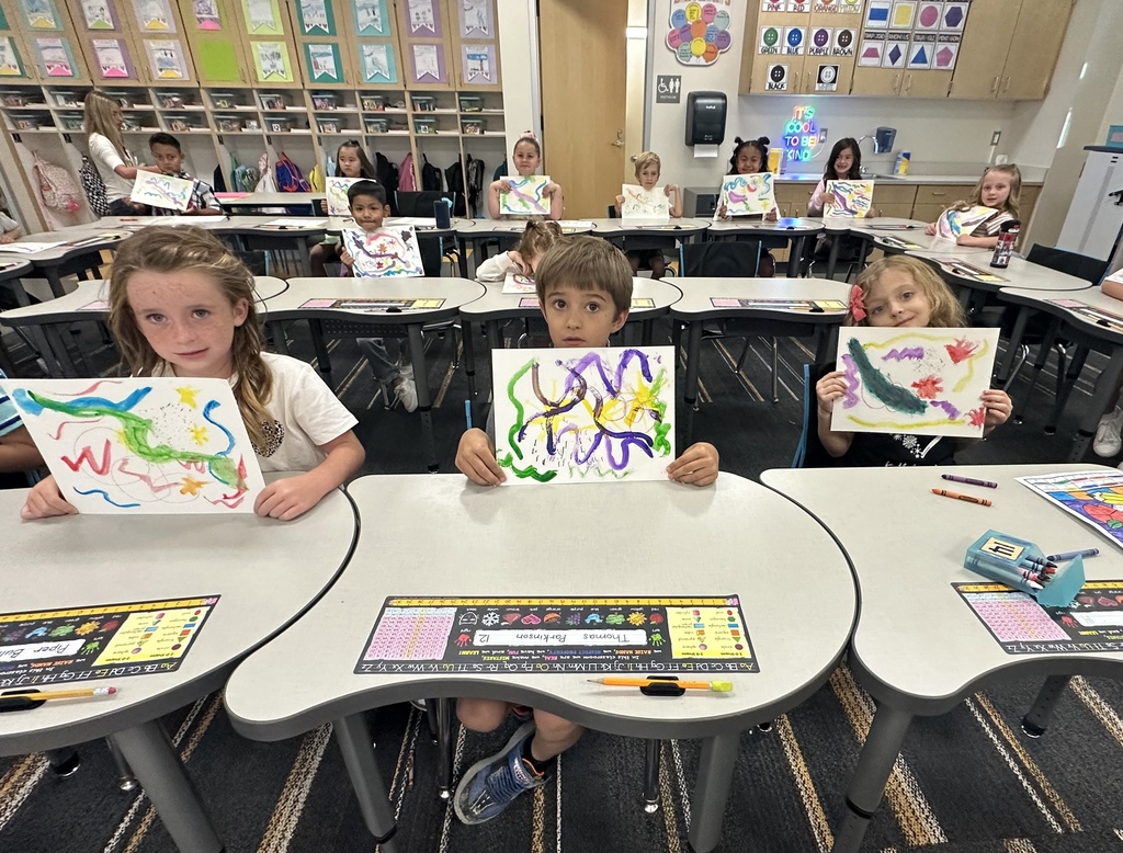 Children at tables in a classroom, each holding up an artwork, surrounded by supplies and other children.