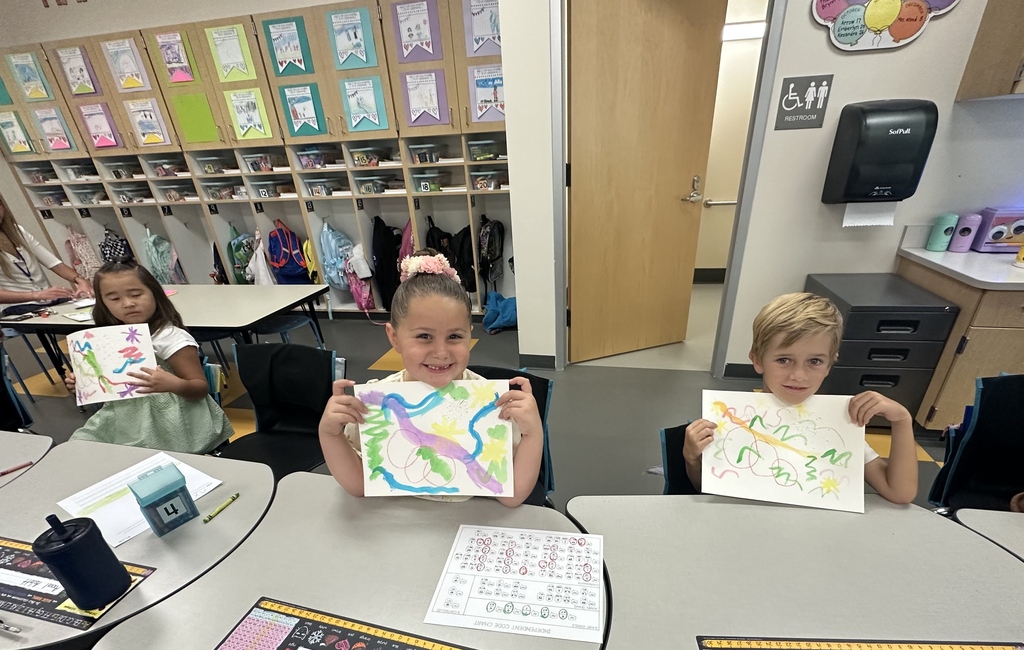 Three children seated at tables in a classroom, holding up colorful drawings. Background includes shelves and cabinets.