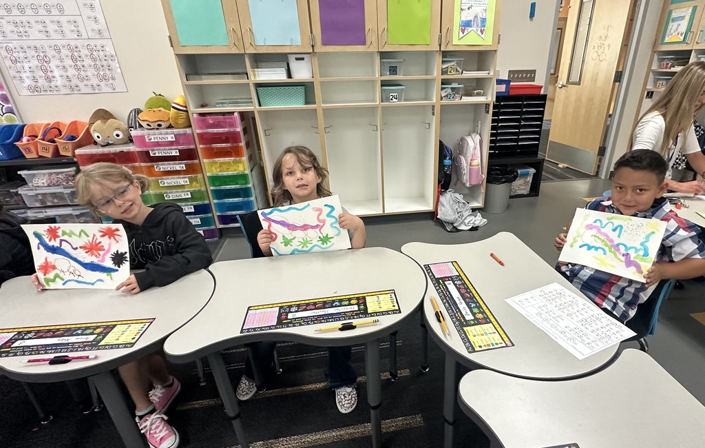 Four children sit at tables in a classroom, holding up colorful drawings. Shelves with various materials and a door are in the background.
