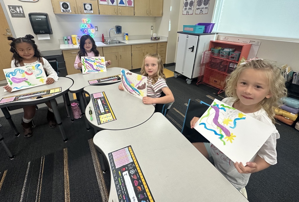 Four girls in a classroom sit at tables with art projects, smiling and holding their drawings.