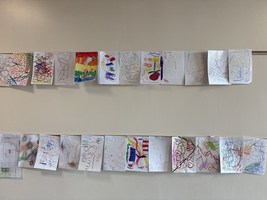 Two rows of children's drawings, varying in size and style, are displayed on a white wall.