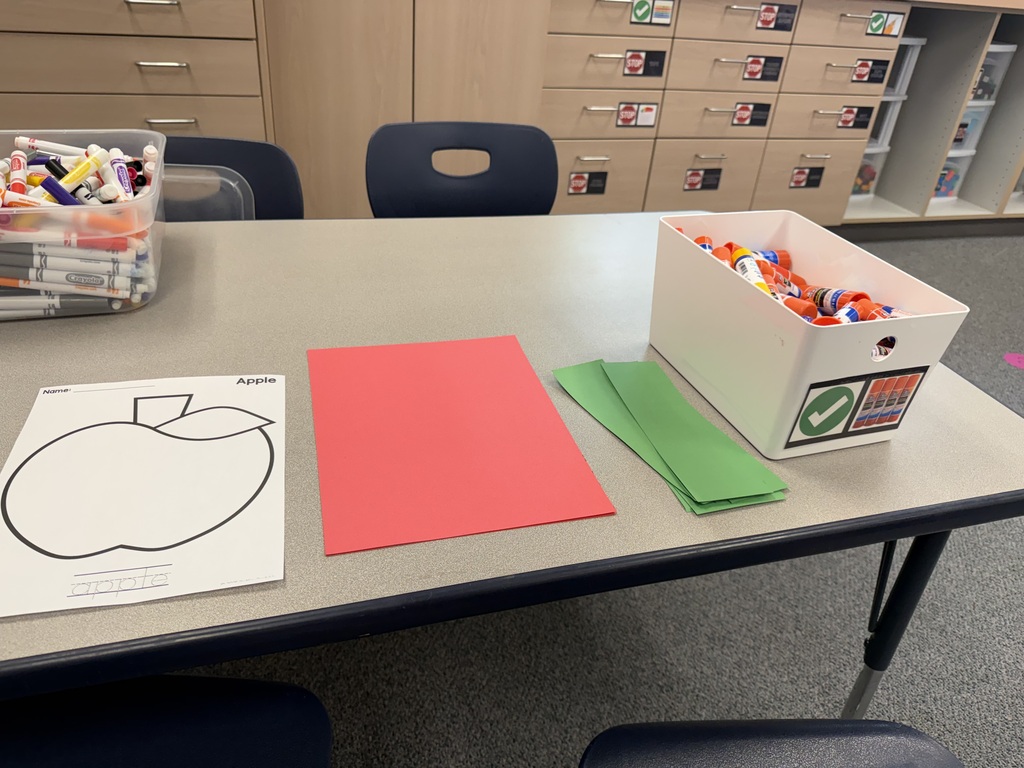 A table displays a box with colorful candies, a red paper apple template, and green paper strips.