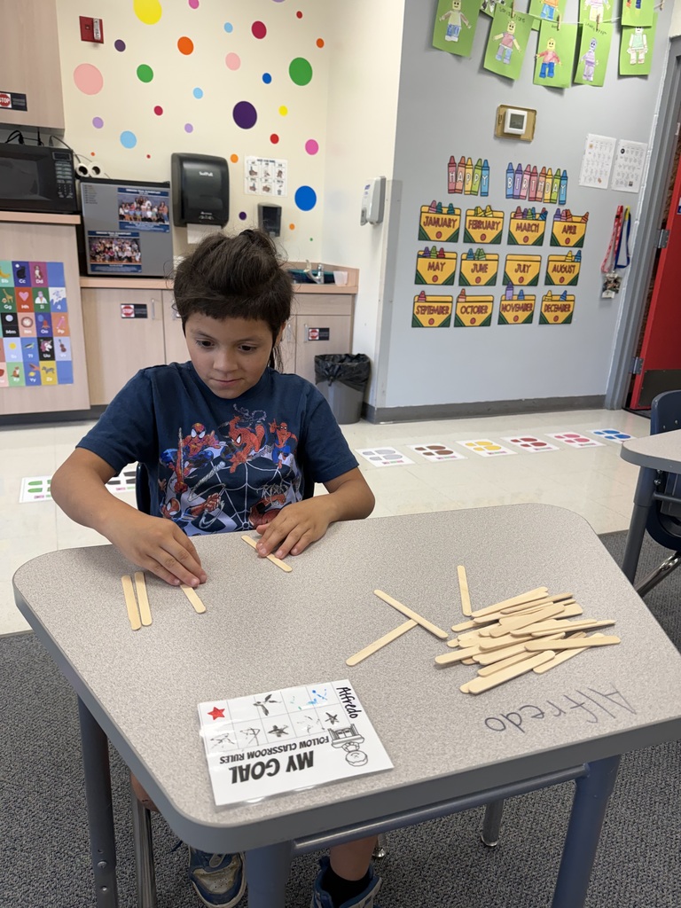 A child sits at a table, using sticks to count a number on a worksheet. The table has colorful dots on the wall in the background.