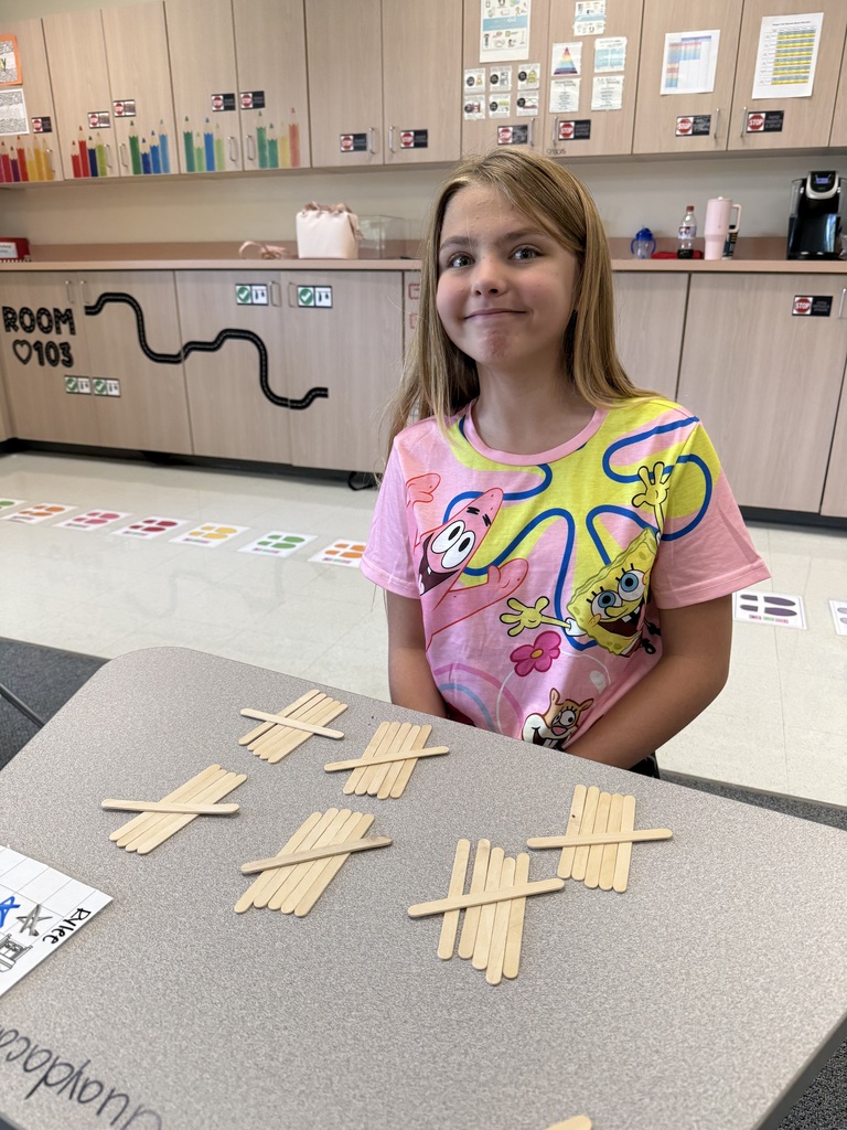 A young girl with blond hair stands at a table in a classroom. She is surrounded by wooden sticks and smiling.