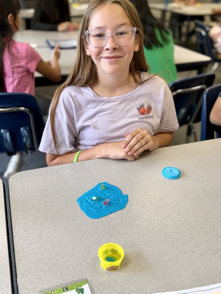 A young girl wearing glasses smiles while sitting at a table with blue and yellow items.
