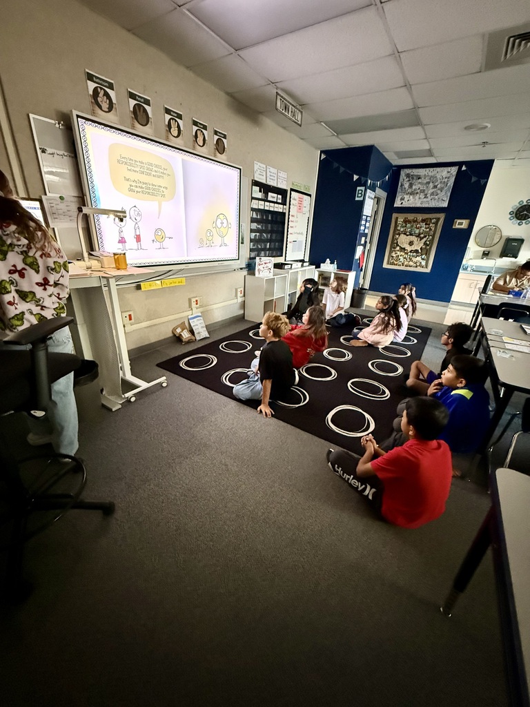 Children sit on a rug in a classroom, facing a large screen displaying an image, while a woman stands near a table.