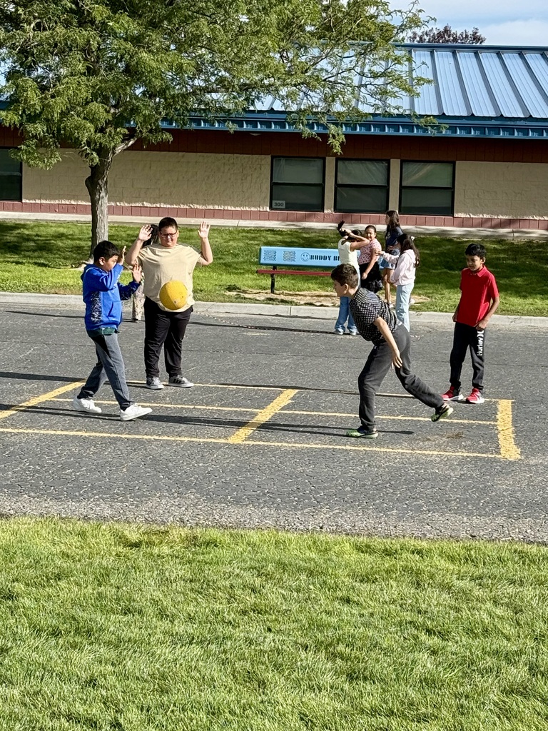 Children play a ball game in a parking lot, surrounded by grass and a building. One child holds a ball.