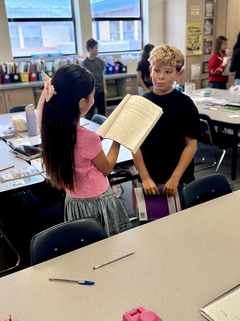 Two kids in a classroom; one holds a book while the other reads aloud. Tables, chairs, and other students in the background.