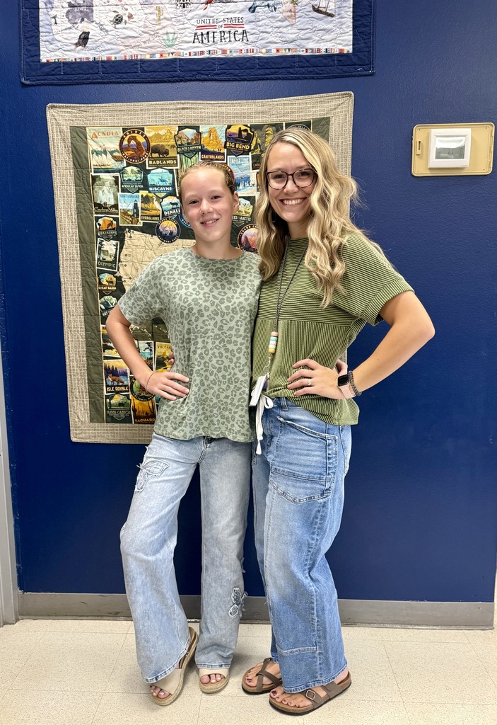 Two young women in green shirts and jeans stand next to each other in front of a blue wall with a quilt.
