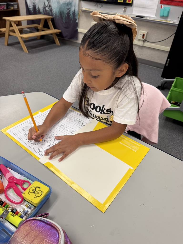 A child in a white shirt writes on a desk with a pencil. A yellow folder with paper sits in front.