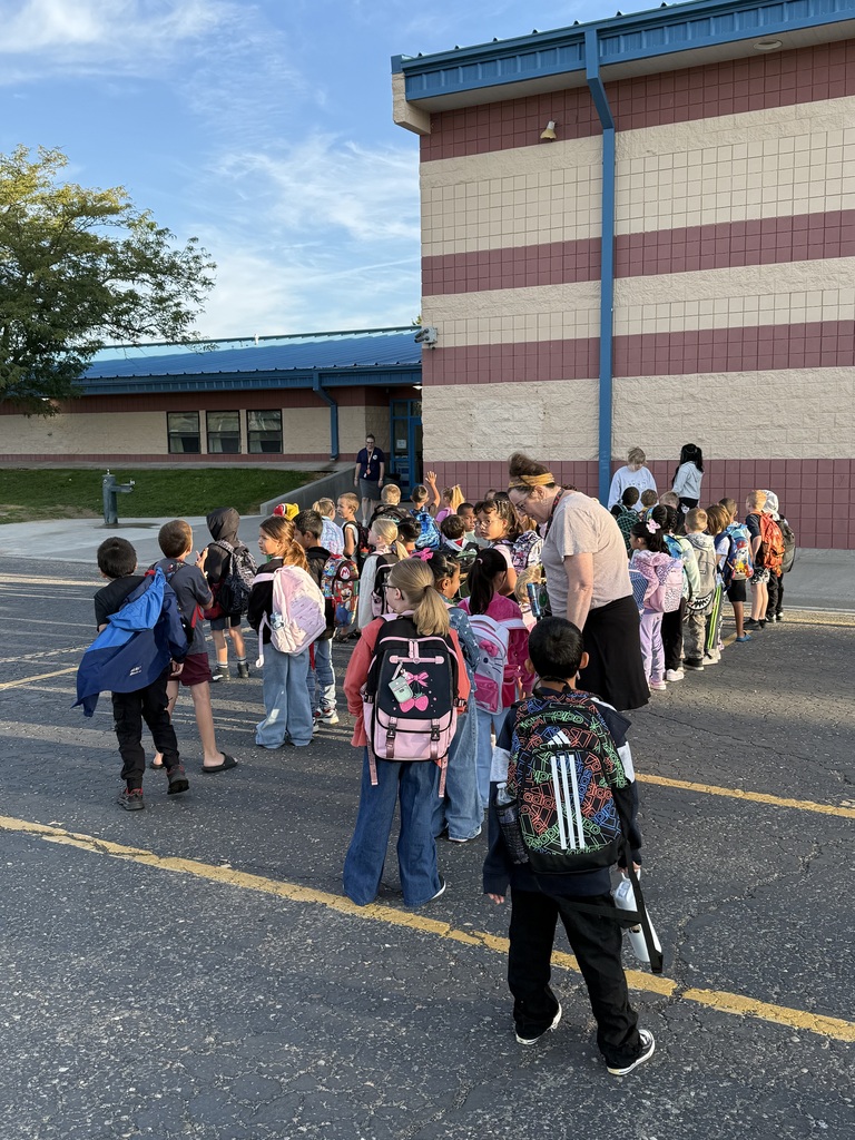 Kids wearing backpacks line up in front of a school building with striped walls and a blue roof.